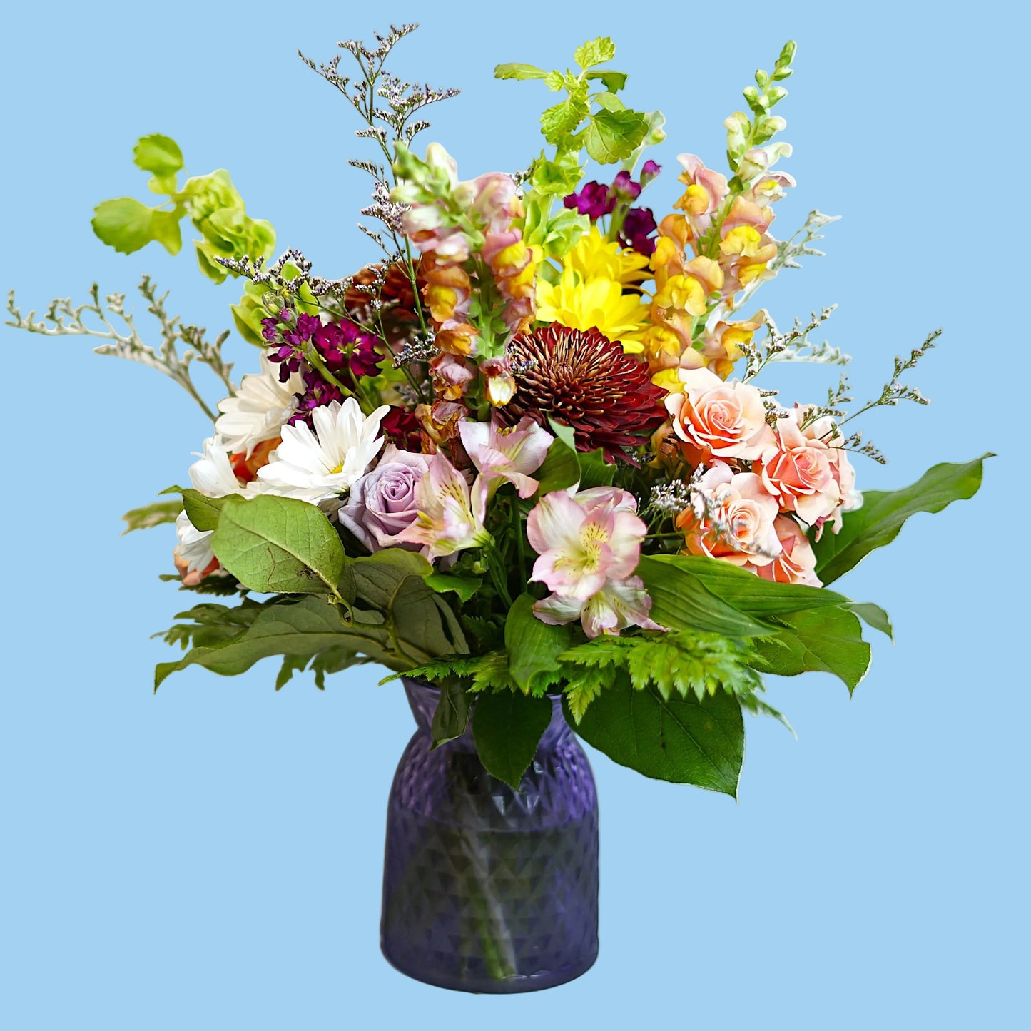 A vibrant mixed floral arrangement in a textured purple glass vase, featuring pink, yellow, white, and purple flowers with lush green foliage, set against a plain light background.